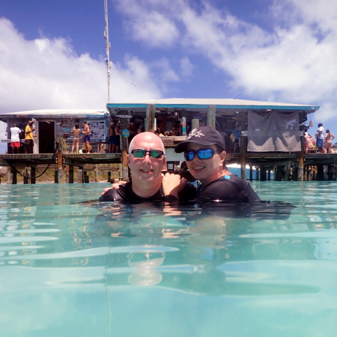 Two people in water with sunglasses, dock and people in the background