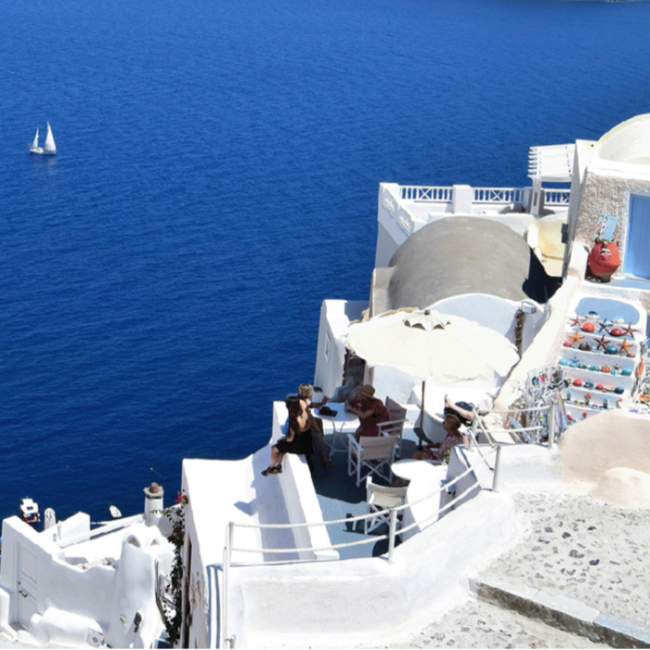 White buildings with blue accents by the sea, with a sailboat in the distance.