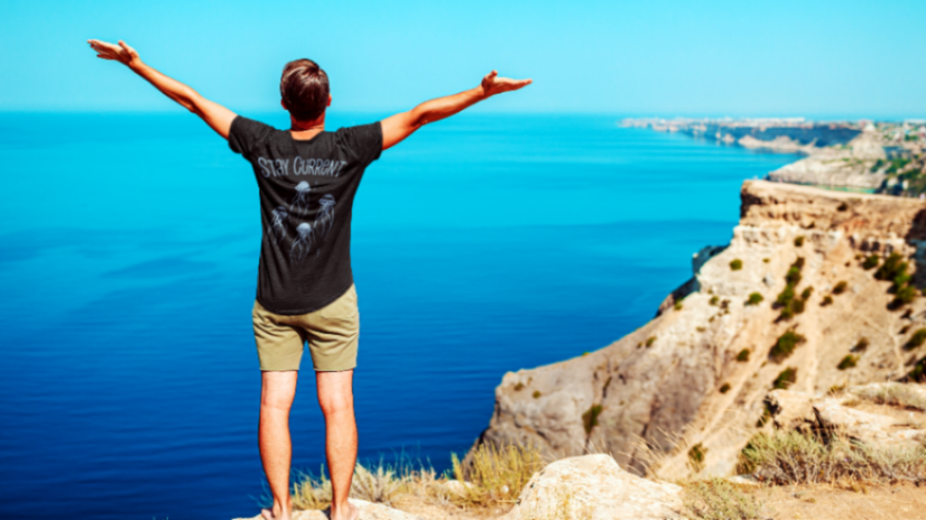 Person standing on a cliff overlooking a blue ocean with arms outstretched.